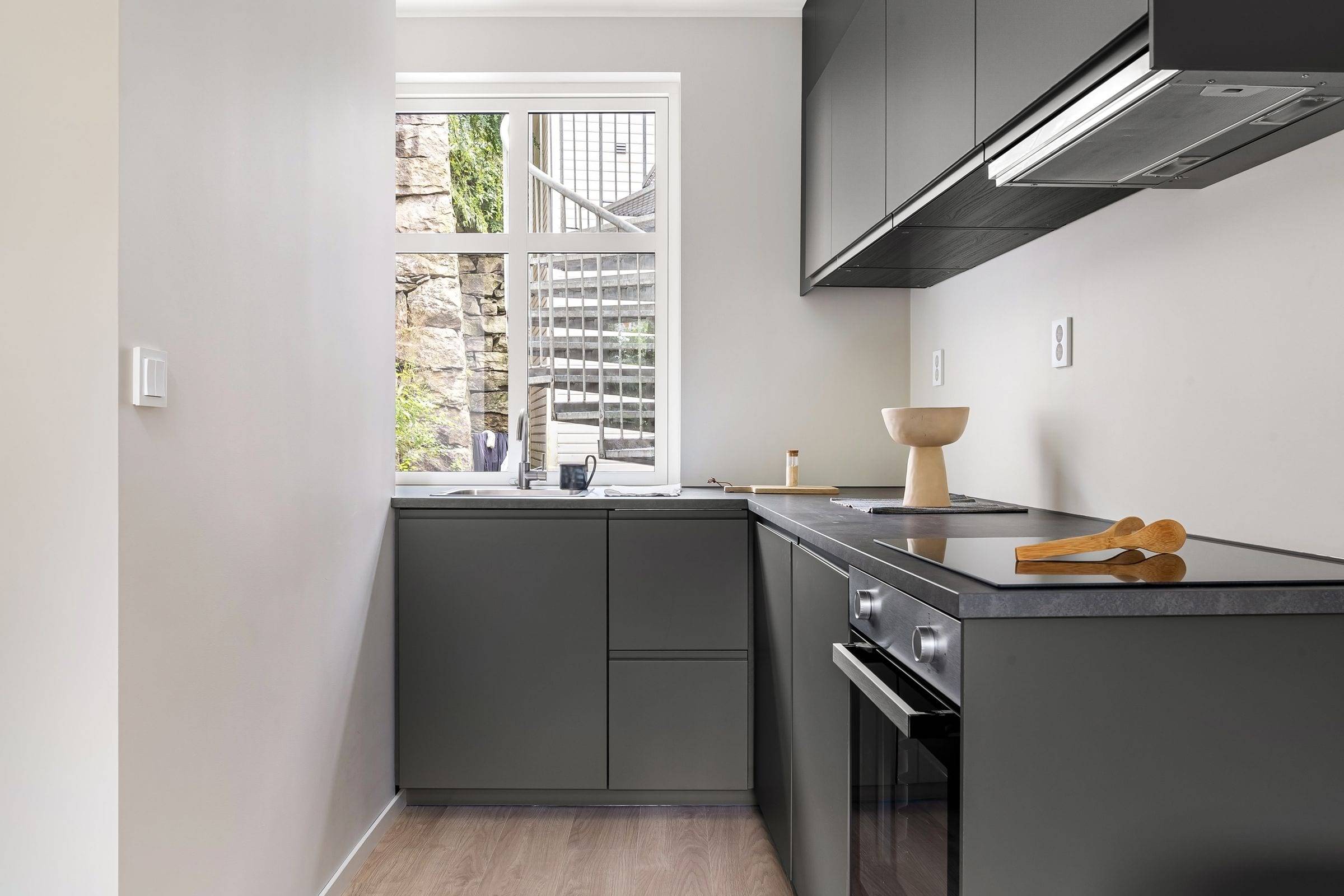 Petite cuisine en L aux façades anthracite mates, plan de travail sombre, évier sous la fenêtre