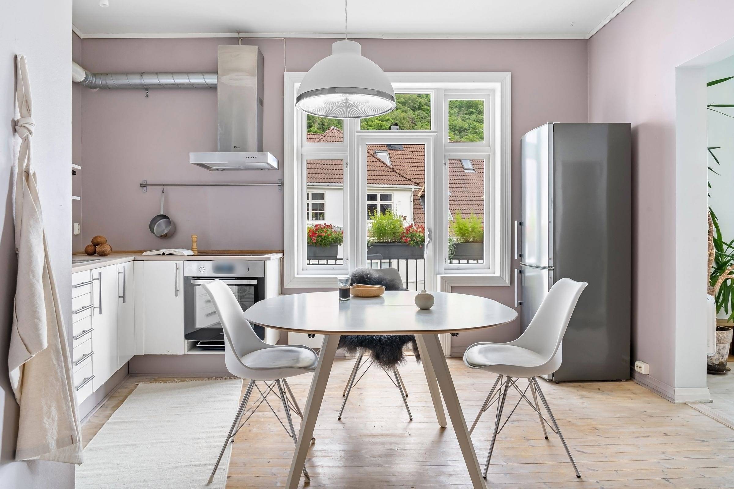 Cuisine en L blanche avec plan de travail en bois, hotte inox et table ronde devant une porte-fenêtre à petits bois