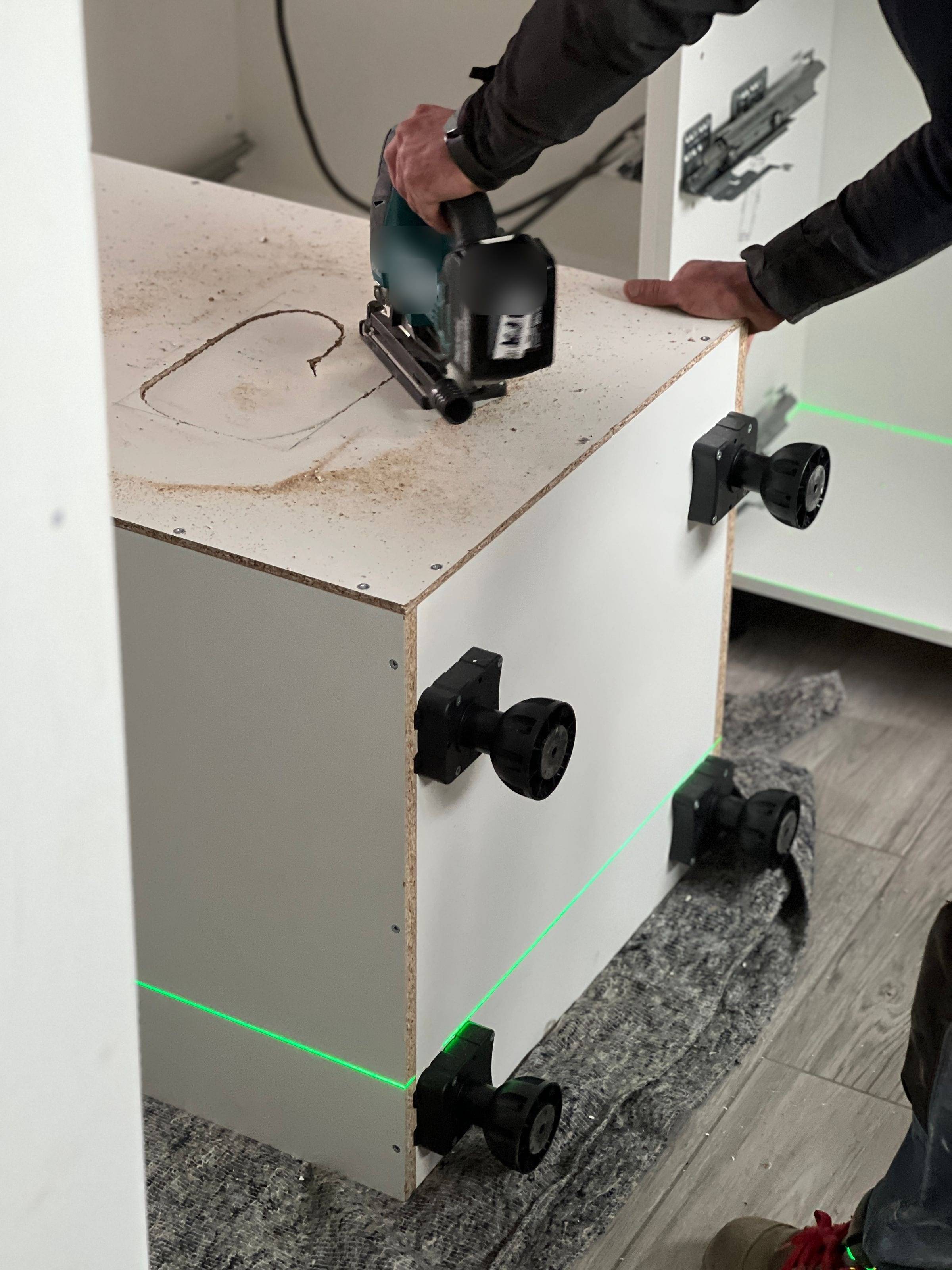 Découpe d’un caisson de cuisine blanc calé sur ses vérins, avec une ligne de niveau laser